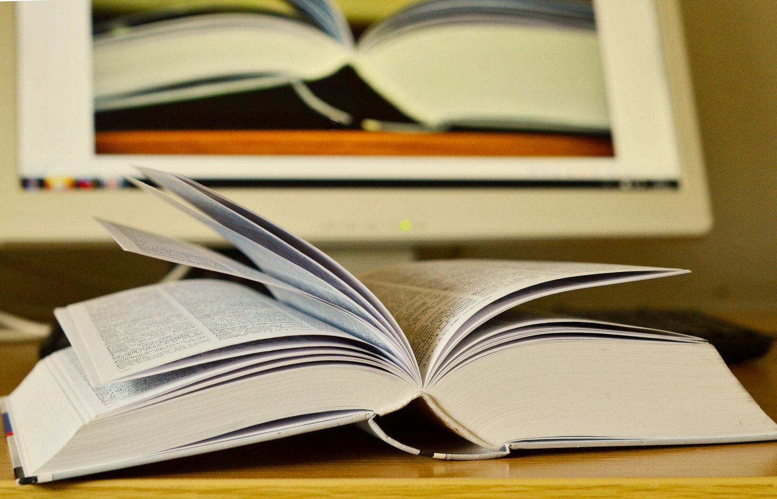 Libro abierto sobre un escritorio, frente a un monitor que muestra la imagen de otro libro abierto